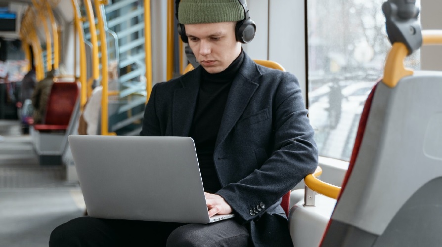 Ung man i sitter med en laptop i knät på ett tåg eller buss.