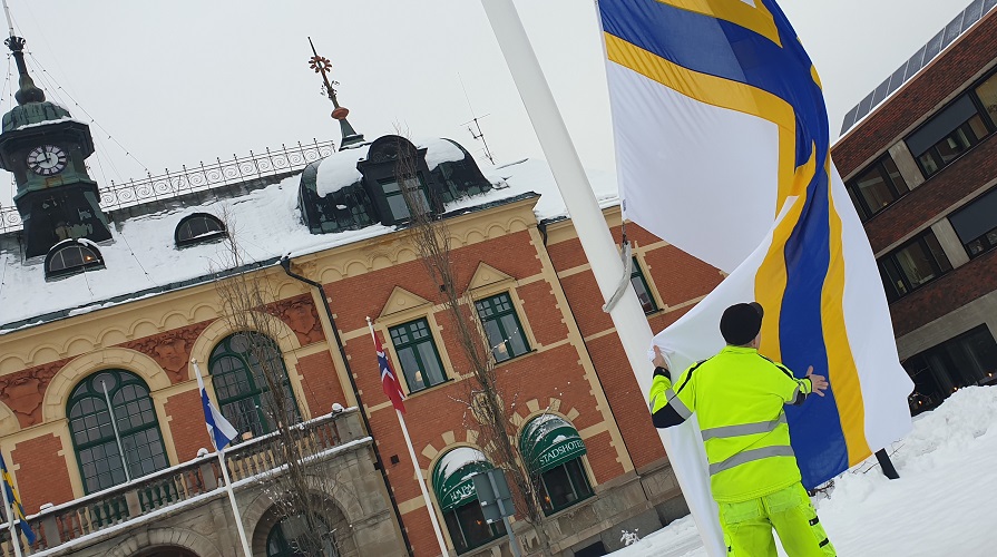 En vit flagga med ett gult och blått kors är på väg att hissas på en flaggstång vid ett torg.