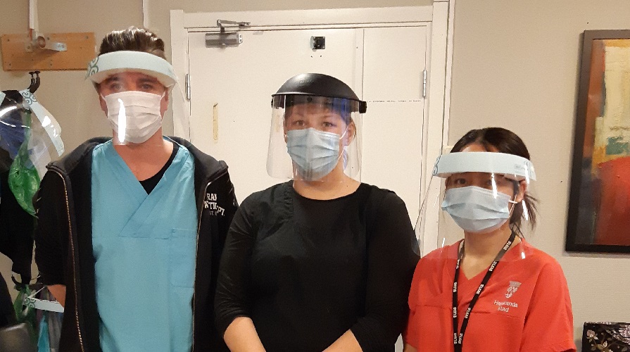 Tre vårdpersonal i mask och visir poserar framför kameran.
