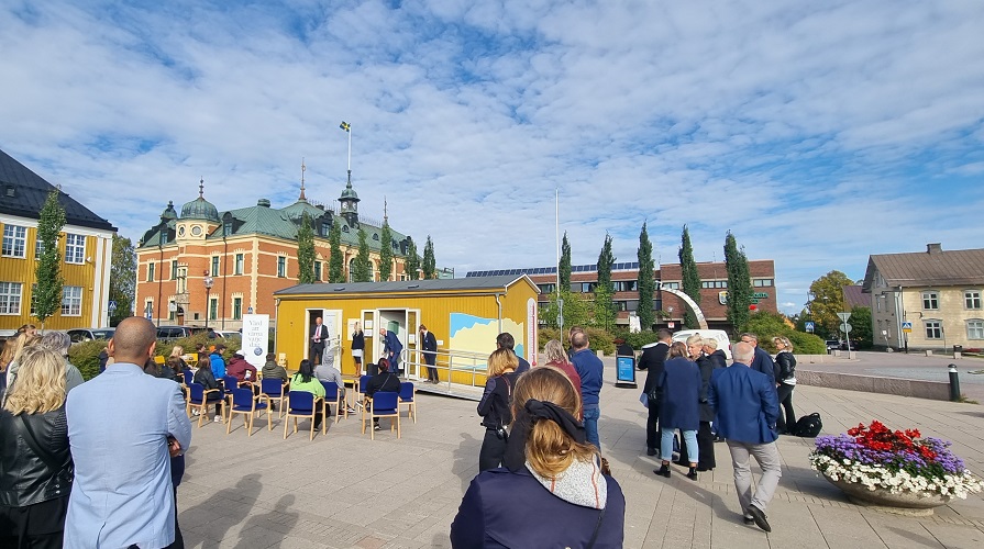 Folksamling på ett torg framför en gul stuga en vacker sensommardag.