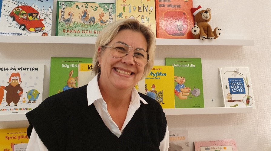 Leende medelålders kvinna i glasögon framför en bokhylla med barnböcker