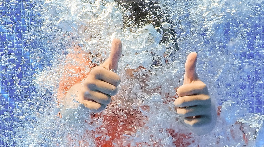 Barn gör tummen upp under vattnet i en pool. 