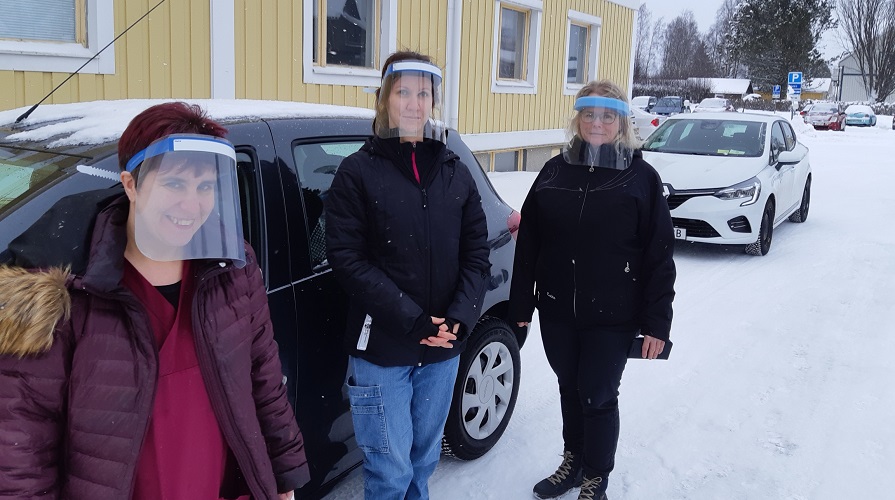 Tre kvinnor med visir poserar framför en byggnad och en bil i ett vinterlandskap.