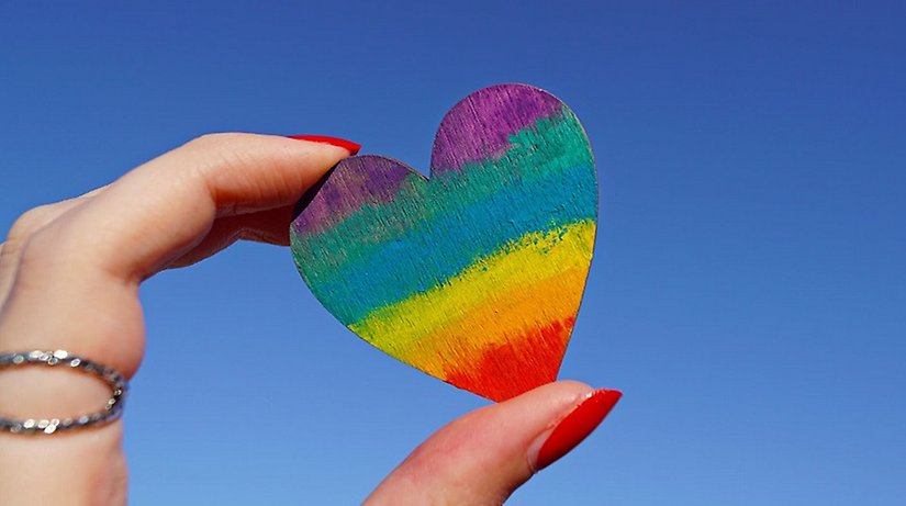 Regnbågsfärgat hjärta hålls upp mot himlen.