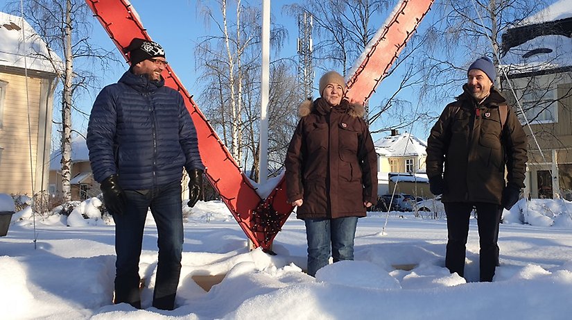 Tre personer på rad poserar på ett snötäckt torg. 