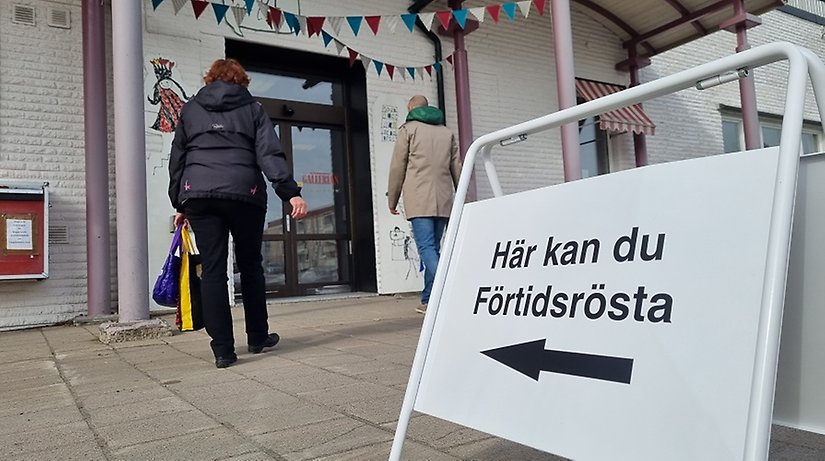 Två personer är på väg in i en lokal. Framför ingången finns en skylt där det står "Här kan du förtidsrösta".