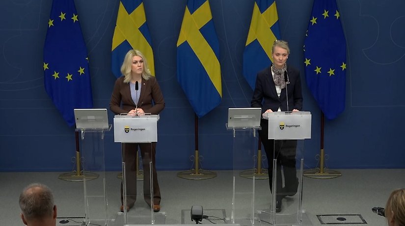 Två medelålders kvinnor på en scen håller presskonferens mot en fond av svenska flaggor och EU-flaggor.