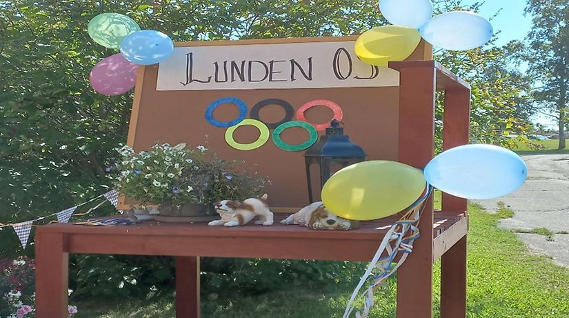 En skylt som visar de fem OS-ringarna samt text Lunden OS. Skylten är omgiven av ballonger och sommarblommor i solens sken. 